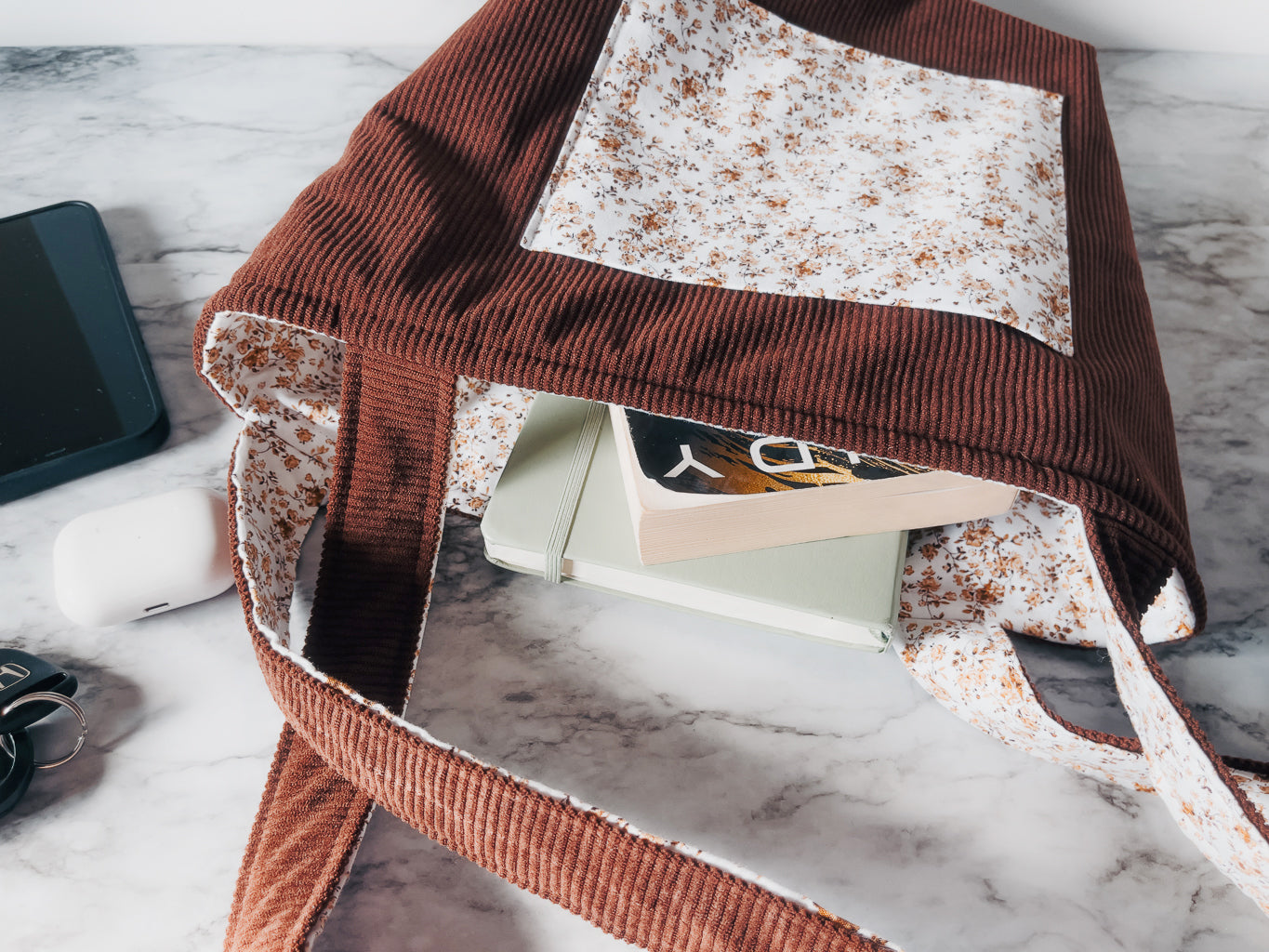 Brown corduroy textured mini tote with front floral slip pocket draped over a marble surface with a phone and mouse visible.