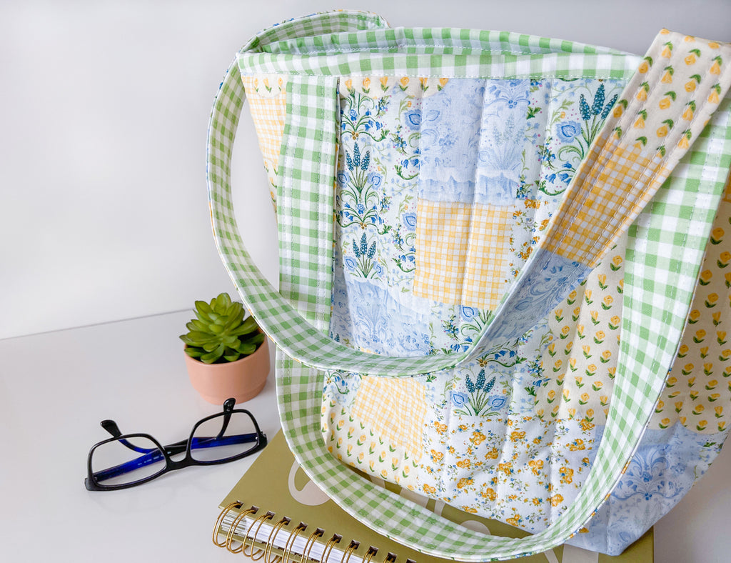 Colorful patchwork quilt with green checkered border on a white surface with glasses and plant.