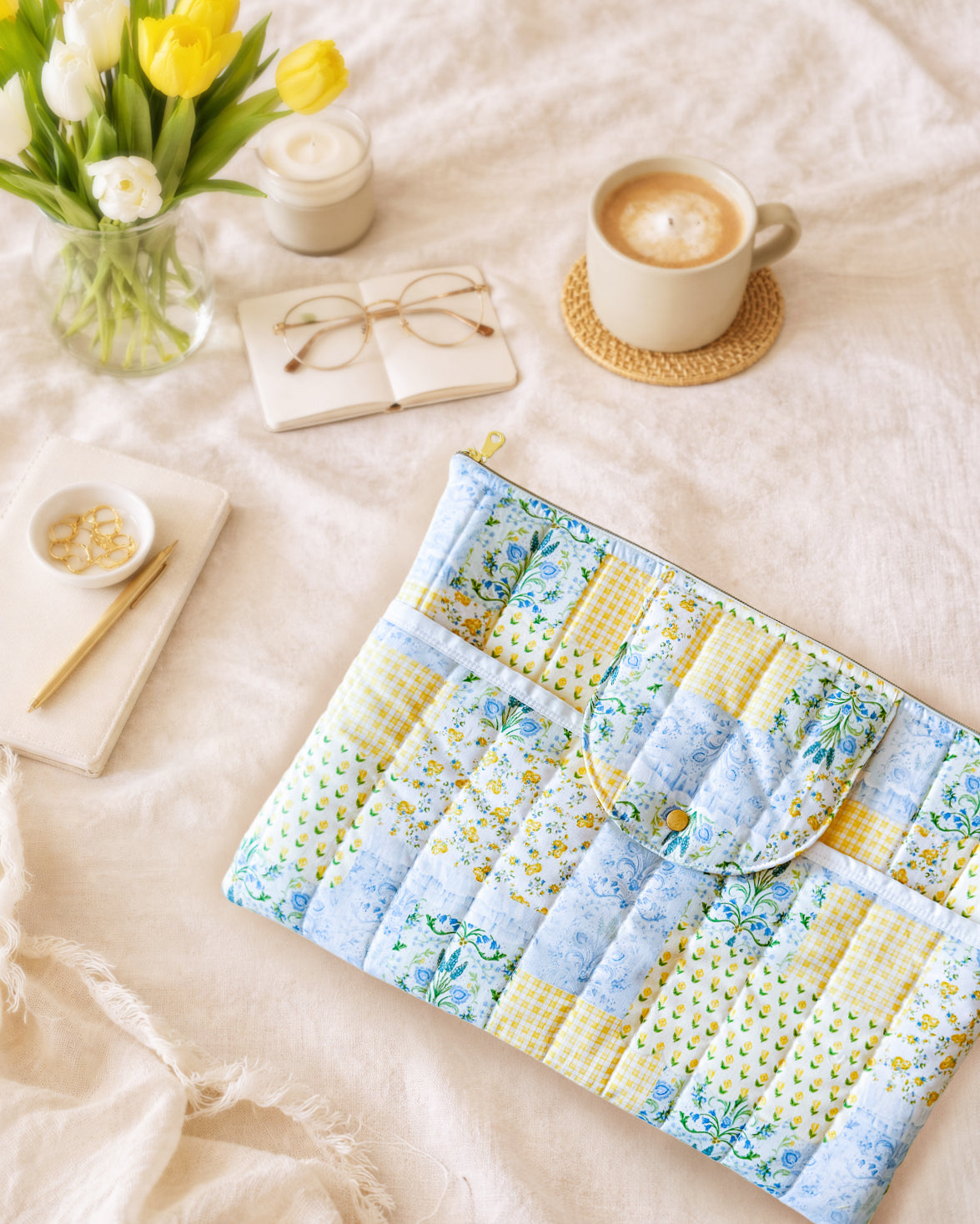 Floral quilted patchwork pattern laptop sleeve on a bed with a cup of coffee, flowers, and candles.